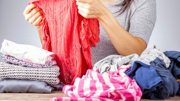 A woman is folding clean dresses on a wooden desk. There is a pile of unfolded dresses and some folded and tidied ones next to it. Concept for house wife, chores and housework.