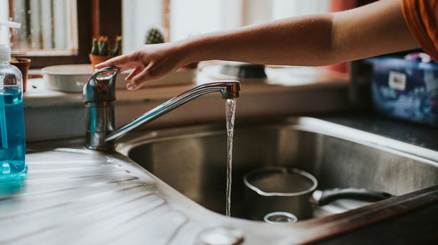 Hand reaching across a sink below a window, to press down tap to turn off the running water. Pot and bowl sit in the sink. Antibacterial hand soap sits off to the side.