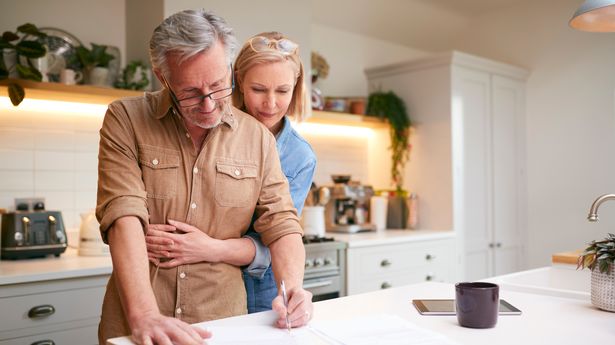 Mature Couple Reviewing And Signing Domestic Finances And Investment Paperwork In Kitchen At Home