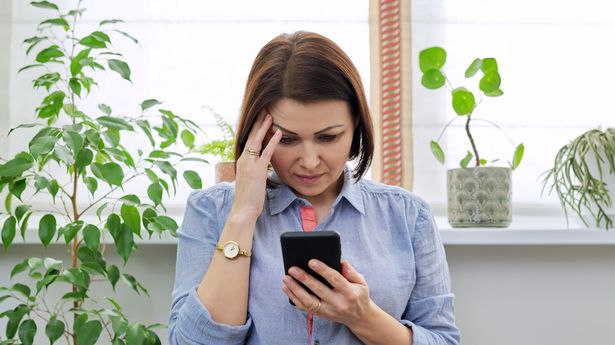 Sad middle aged woman with phone at home near the window