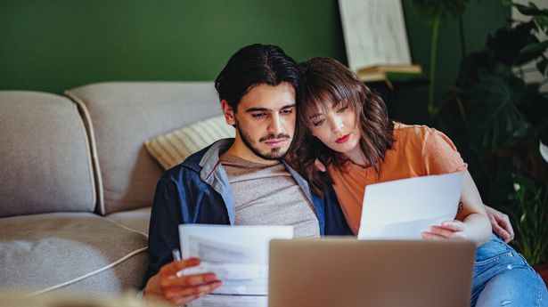 Boyfriend and girlfriend holding bills and doing home finances together online on a laptop computer in the living room.