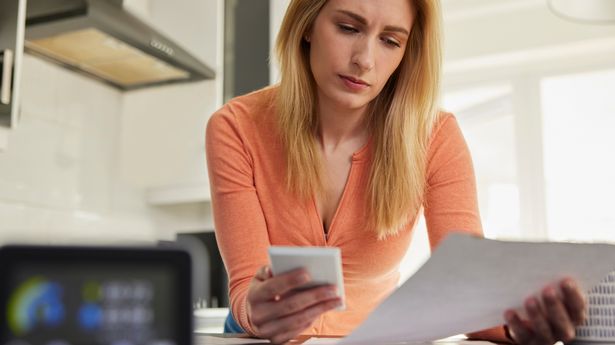 Smart Energy Meter In Kitchen Measuring Electricity And Gas Use With Woman Looking At Bills With Calculator