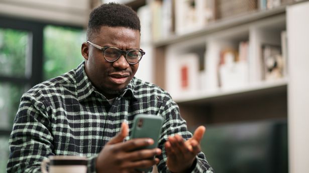 Annoyed man sitting at home, reading some bad news on his smart phone using a mobile app and expressing his displeasure